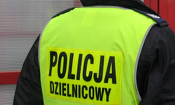 Stojący tyłem policjant z założoną kamizelką odblaskową z napisem Policja Dzielnicowy.